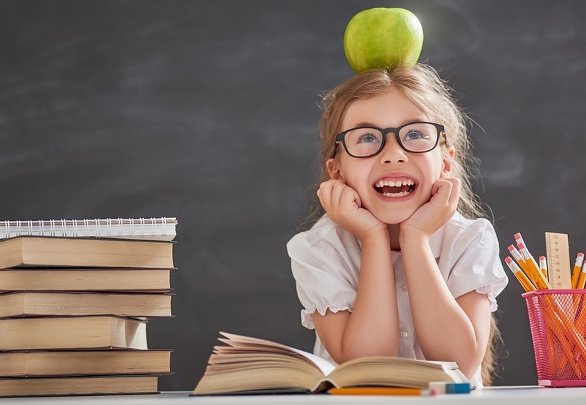 Lerntherapie: Mädchen mit Apfel auf dem Kopf sitzt vor Büchern und freut sich